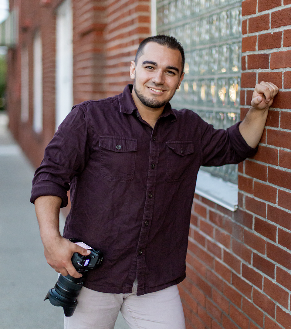 Smiling man in a dark purple shirt holding a camera, leaning against a brick wall with glass block window panels.