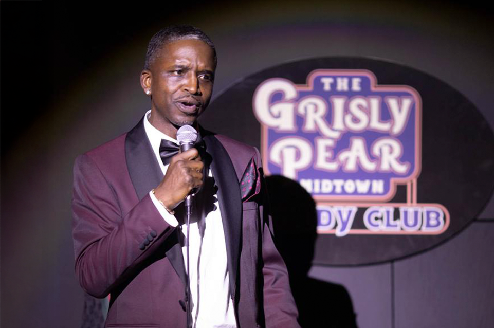 Male comedian in a maroon tuxedo holding a microphone on stage with The Grisly Pear Comedy Club sign in the background.