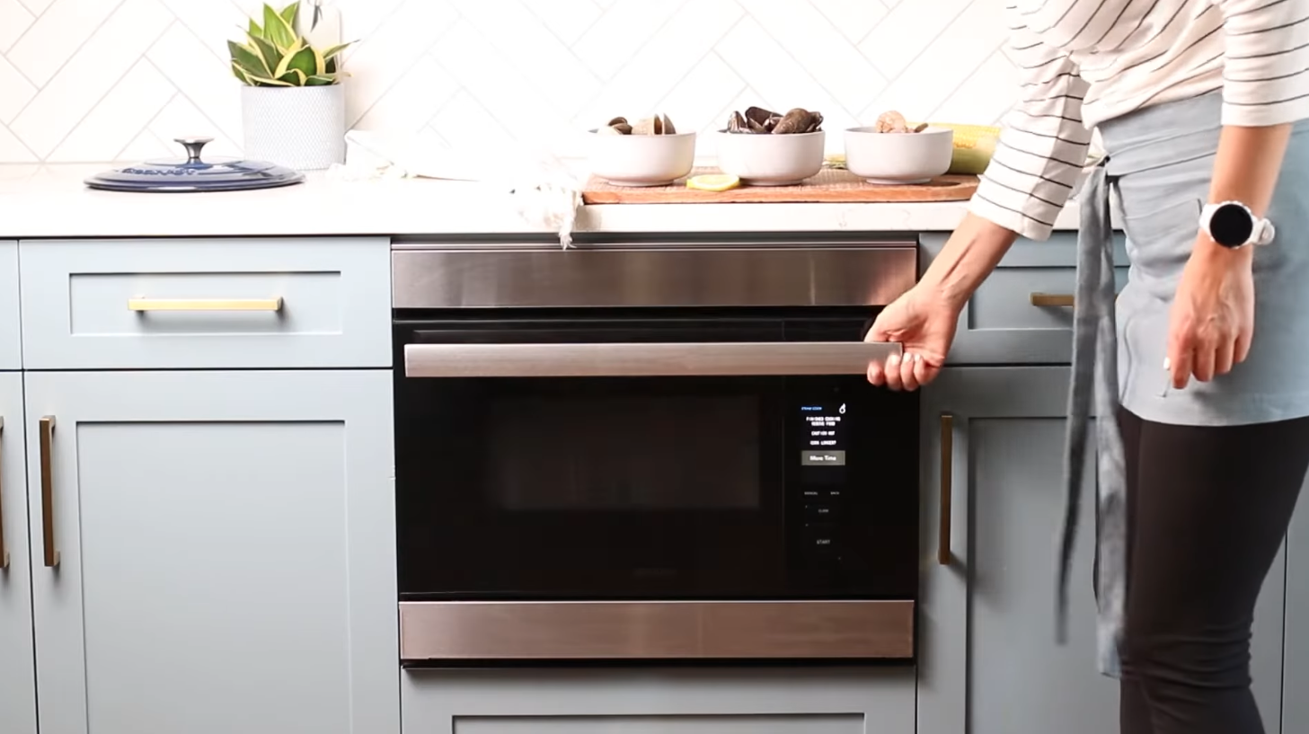 Person opening a built-in stainless steel oven door in a modern kitchen with light blue cabinets and white countertop.