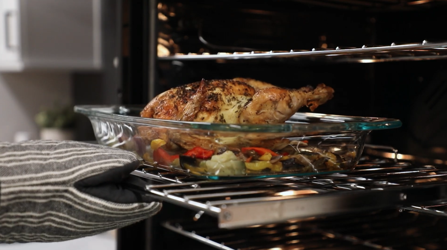 Person wearing a striped oven mitt placing a glass baking dish with a roasted chicken and vegetables into an oven.