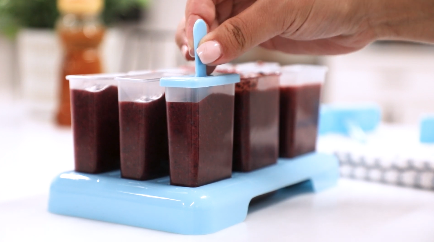 Hand placing a blue lid on a filled popsicle mold with dark purple mixture on a blue stand.