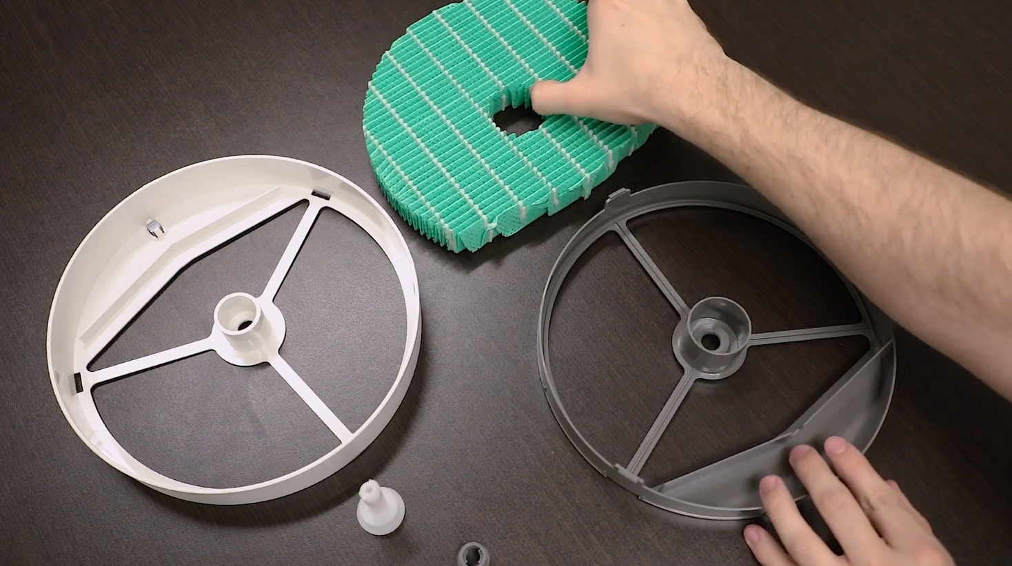 Hands assembling parts of a humidifier filter, including a green pleated filter and plastic circular frames on a dark surface.