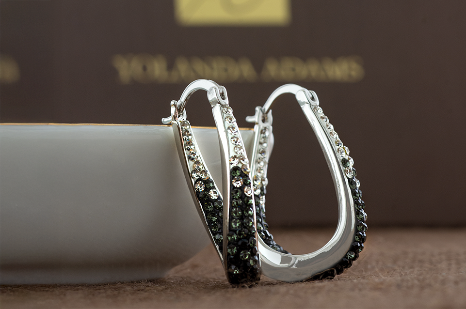 Silver hoop earrings embellished with small black and clear gemstones against a brown background with blurred Yolanda Adams branding.