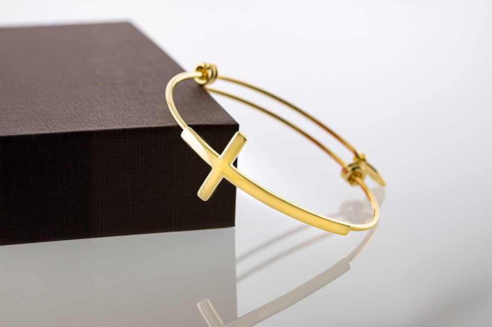 Gold bangle bracelet with a horizontal cross resting on a textured dark brown box.