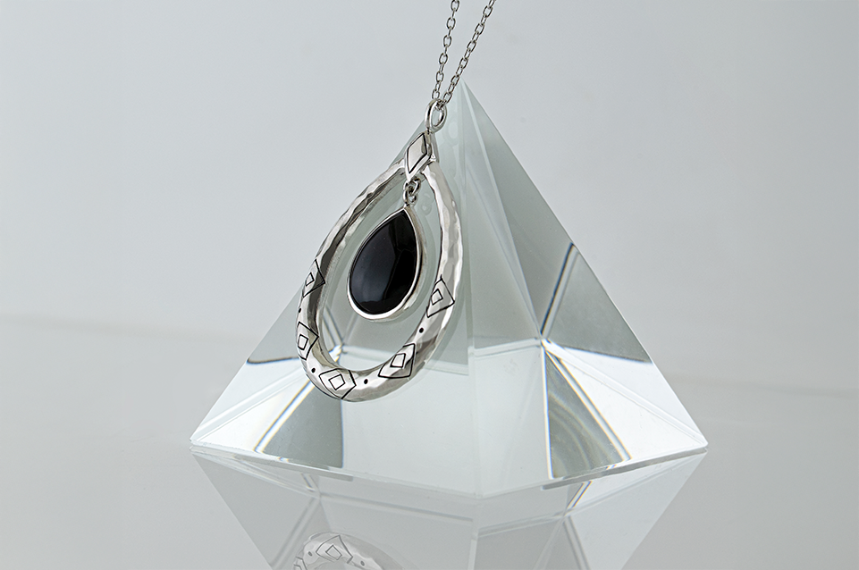 Silver teardrop-shaped pendant with black stone in center hanging on a chain over a clear pyramid-shaped crystal.