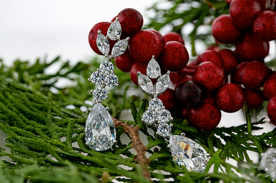 Pair of diamond earrings with a teardrop shape at the bottom, displayed on green pine leaves with red berries in the background.