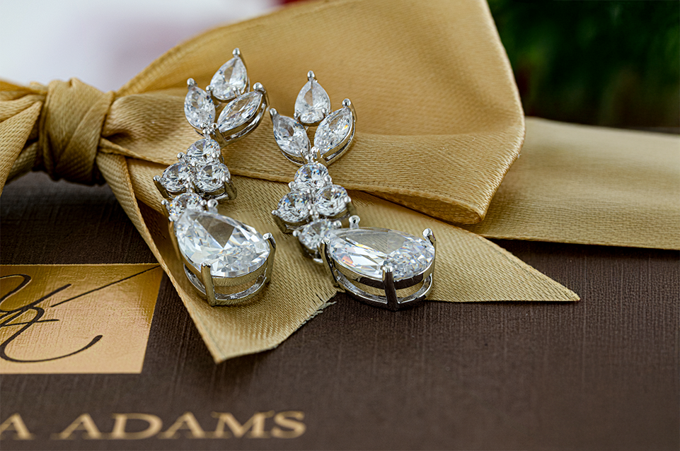 Pair of silver drop earrings with clear teardrop and marquise-shaped gemstones resting on a golden fabric bow over a brown surface with text.