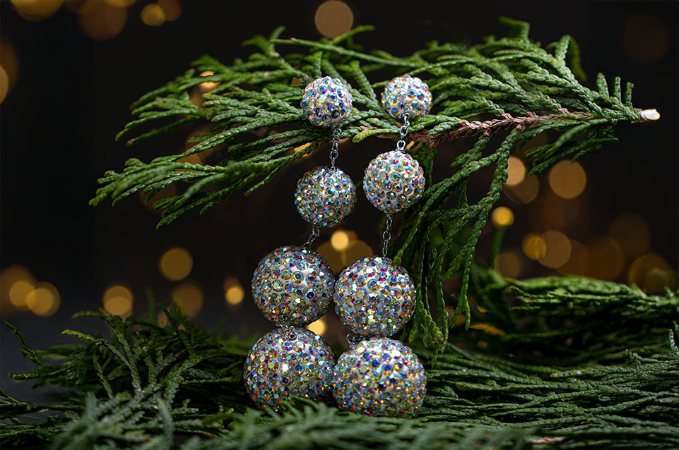 Dangling earrings composed of four connected glittering spherical beads adorned with multicolored rhinestones, displayed among green pine branches with a dark, softly lit background.