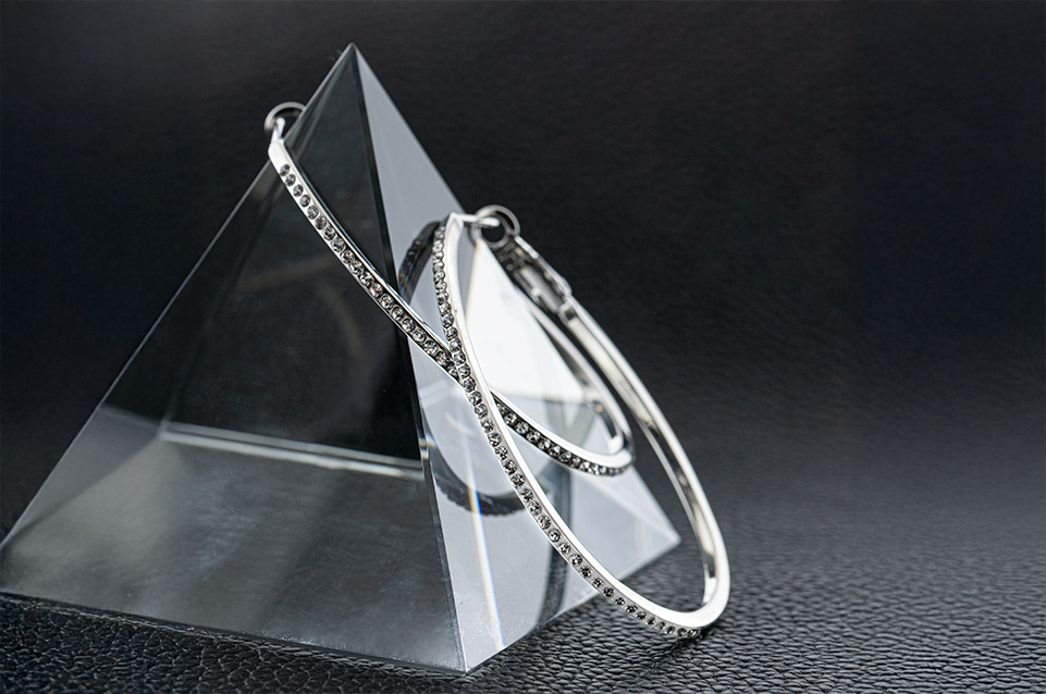 Silver hoop earrings encrusted with small clear stones displayed on a clear pyramid stand over a textured black surface.