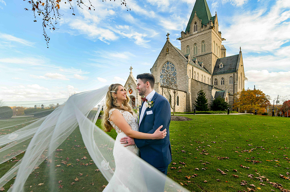 Bride with flowing veil and groom embracing on green lawn in front of large stone church under blue sky.