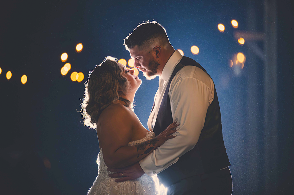 Bride and groom embracing closely, illuminated from behind against a dark background with warm bokeh lights.