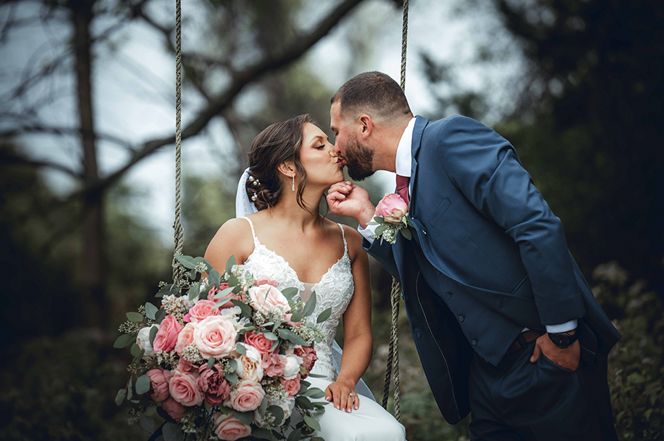 Bride in white dress holding a pink rose bouquet kisses groom in blue suit who gently holds her chin outdoors.