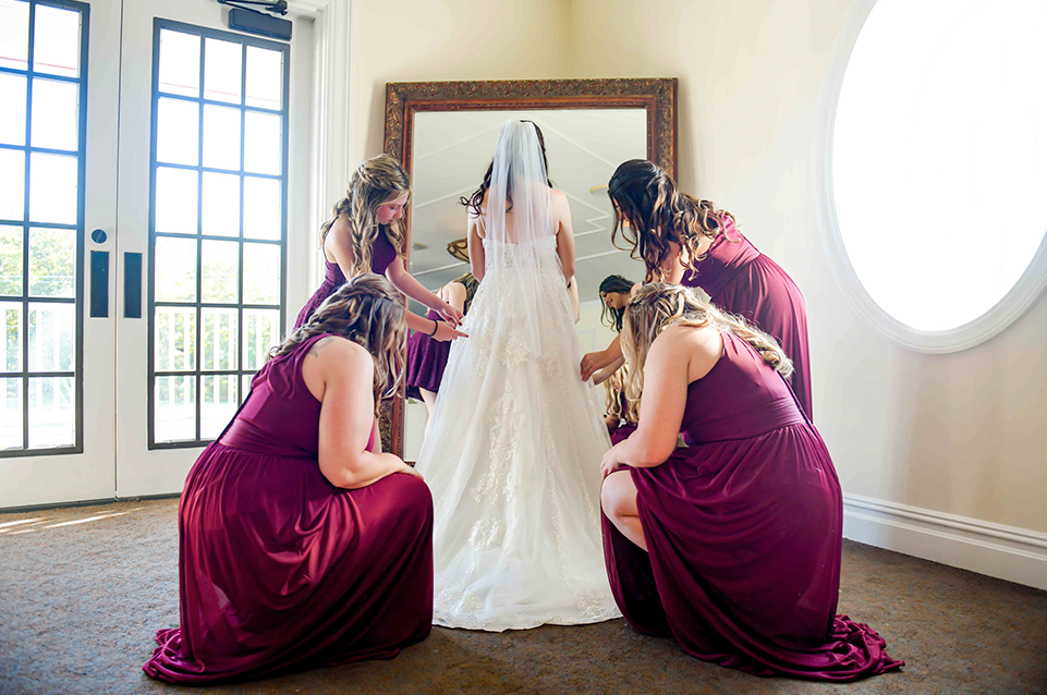 Bride wearing a white wedding dress with veil stands before a large mirror while bridesmaids in matching burgundy dresses adjust her gown.