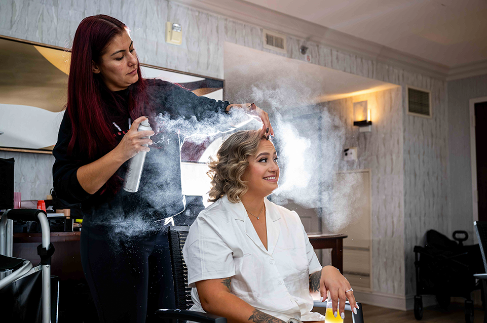 A woman with red hair sprays hairspray on a seated smiling woman with styled wavy blonde hair in a well-lit room.