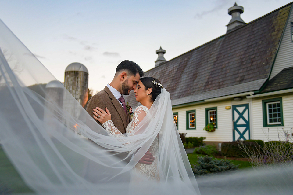 Bride and groom embracing intimately outdoors with bride’s veil flowing, in front of a rustic barn at dusk.