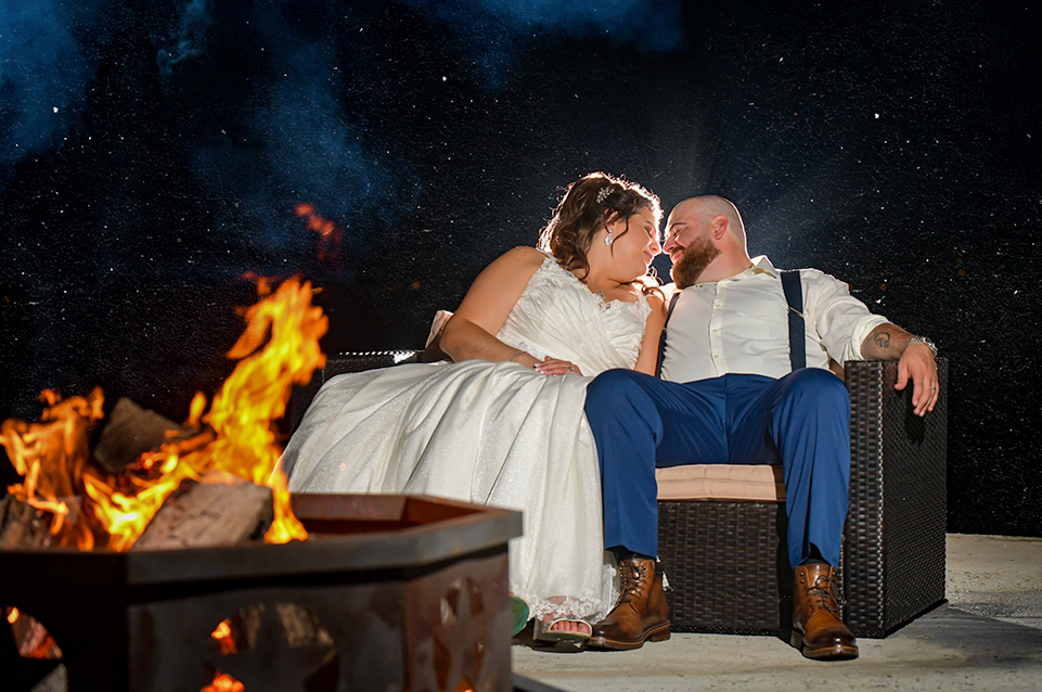 Bride and groom sitting closely on a wicker bench by a fire pit at night, looking lovingly at each other.
