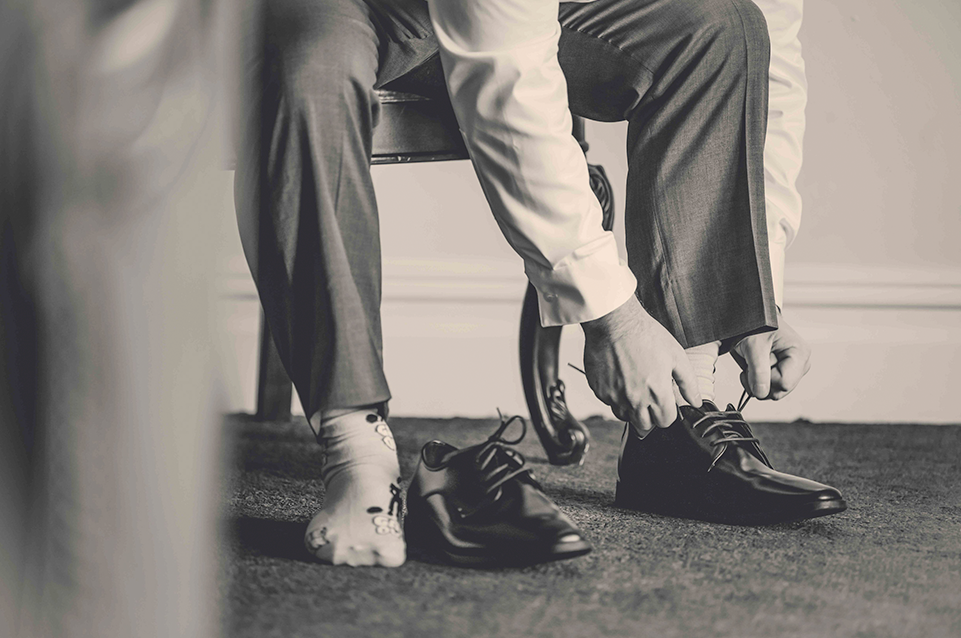 Man sitting on a chair tying the laces of one black dress shoe while wearing a sock with cartoon characters on the other foot.