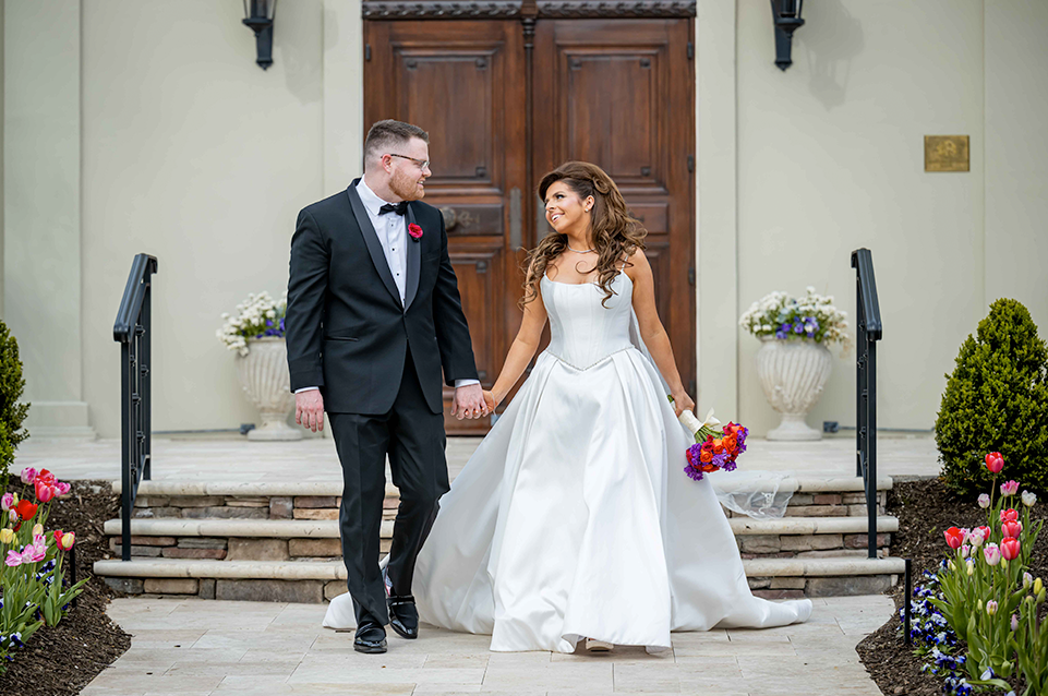 Bride in a white dress holding a bouquet and groom in a black tuxedo walking hand-in-hand outside a building with wooden doors and flowerbeds.