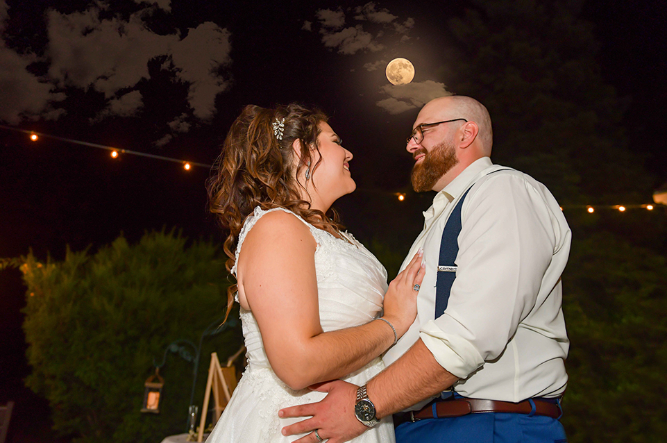 Bride and groom in wedding attire embracing and smiling under a night sky with a full moon and string lights.