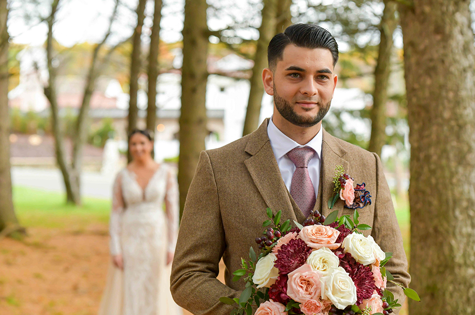 Smiling groom in brown suit holding a bouquet of flowers with bride in white wedding dress blurred in the background among trees.