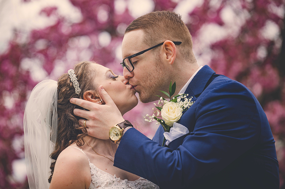 Bride and groom sharing a kiss outdoors with pink blossoming trees in the background.
