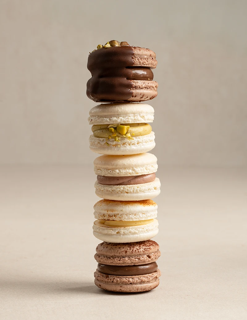 Stack of assorted macarons with pistachio, chocolate, crème brûlée, mixed berry, and Dubai chocolate flavors.