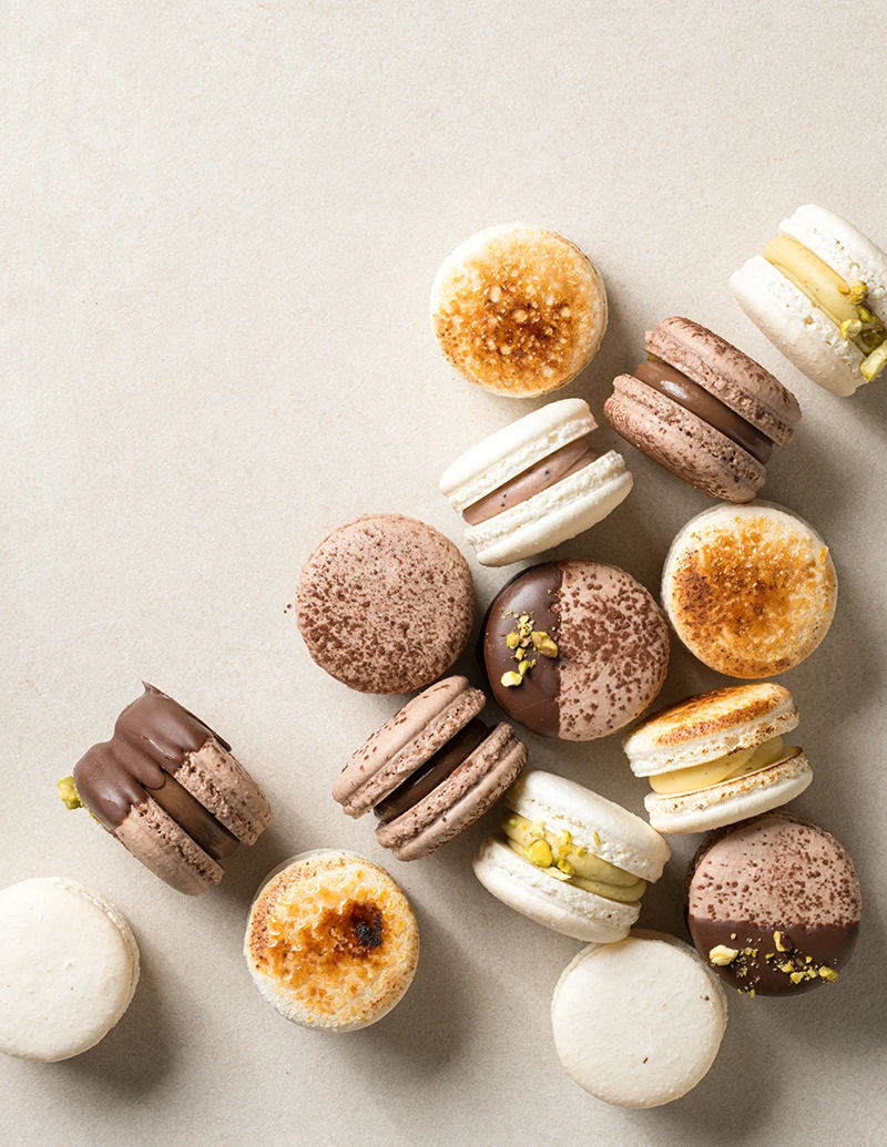 Assorted natural macarons scattered on a neutral surface with pistachio, chocolate, crème brûlée, mixed berry, and Dubai chocolate fillings.