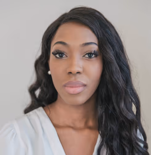 Portrait of a woman with long black wavy hair, wearing pearl earrings and a light-colored blouse, looking directly at the camera.