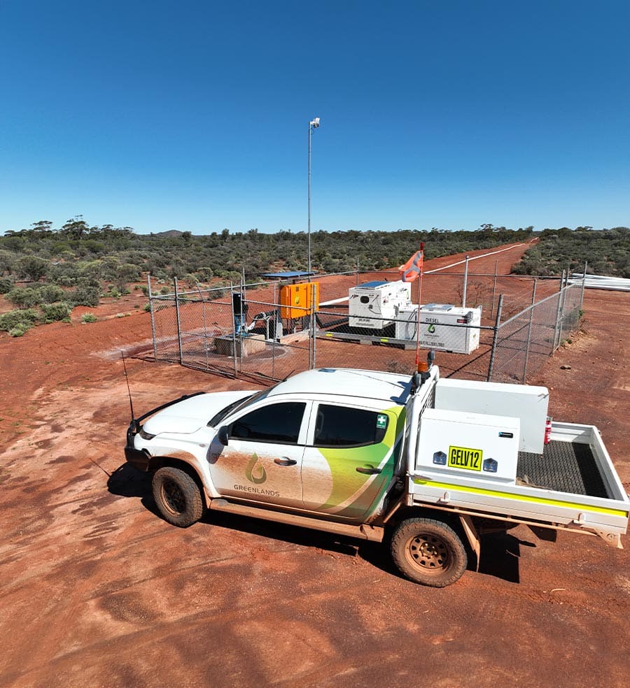 White and green pickup truck with Greenlands logo parked on red dirt near a fenced utility area with equipment under a clear blue sky.