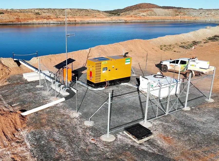 Fenced industrial pump station with yellow generator, pipes, a white equipment container, and a white utility truck near a large water reservoir in an arid landscape.