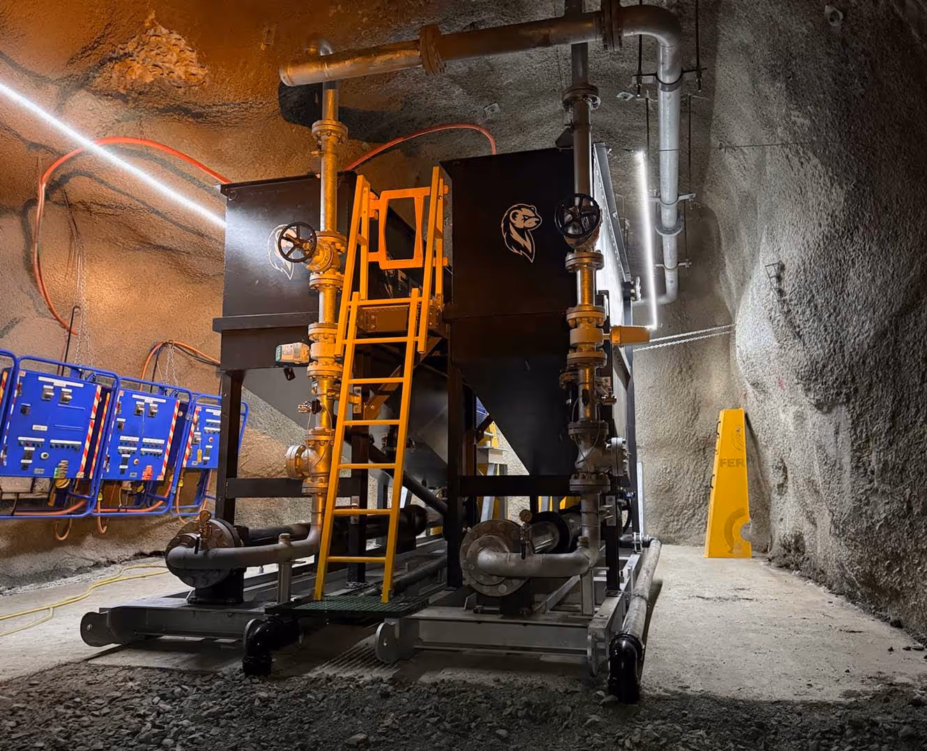 Industrial machinery with pipes, valves, and yellow ladder inside a rocky underground tunnel with blue control panels on the wall.