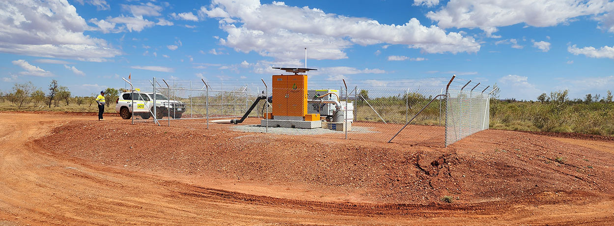Industrial pump connected to white pipes in a red dirt construction area with a pickup truck in the background.