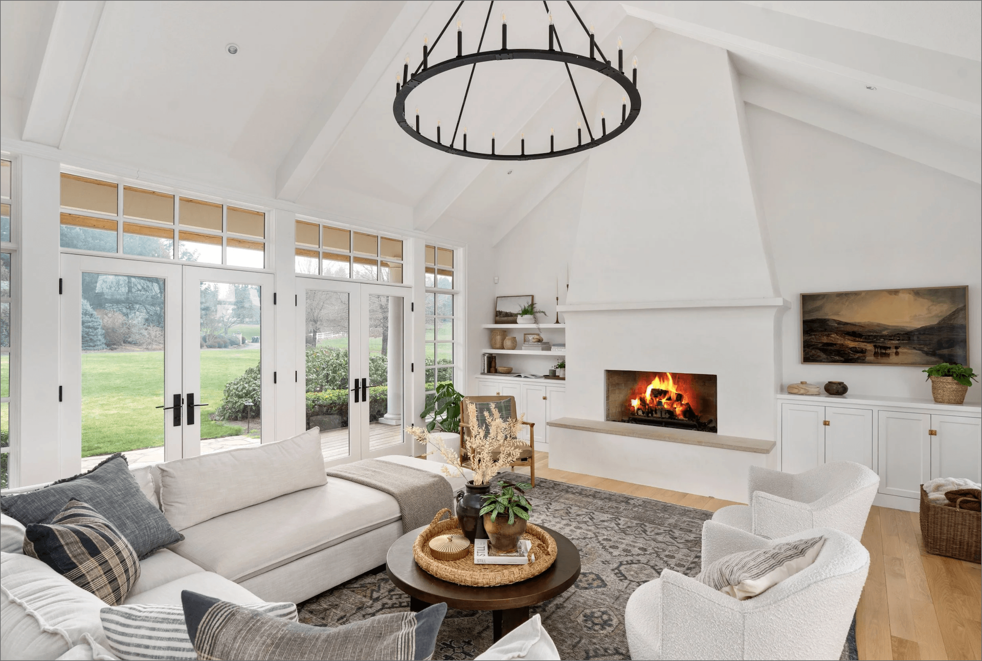 Bright living room with white sectional sofa, round wooden coffee table, two white armchairs, a lit fireplace, large windows looking out to a green lawn, and a circular black chandelier.