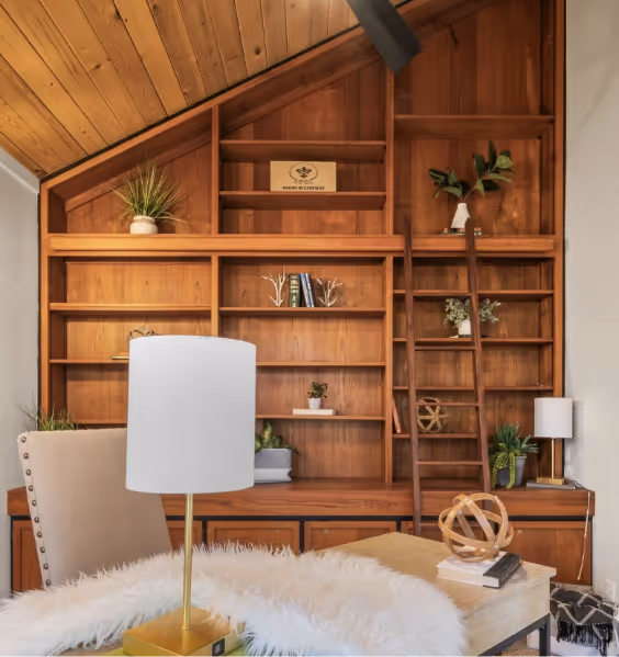 Cozy wooden study room with slanted ceiling, built-in shelves, ladder, beige upholstered chair, and white table lamp.
