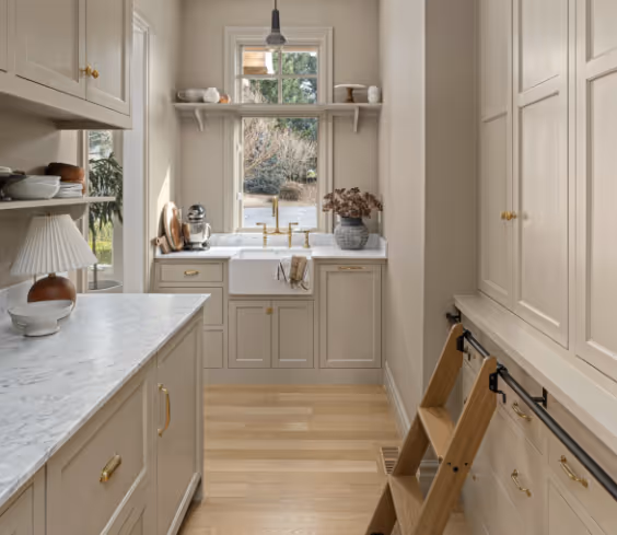 Bright kitchen with beige cabinetry, marble countertops, farmhouse sink, wooden floor, and a wooden step ladder.