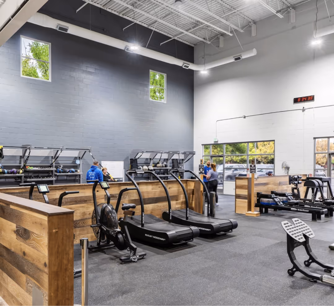 Modern gym interior with wooden partitions, exercise machines, and two people talking near the entrance.