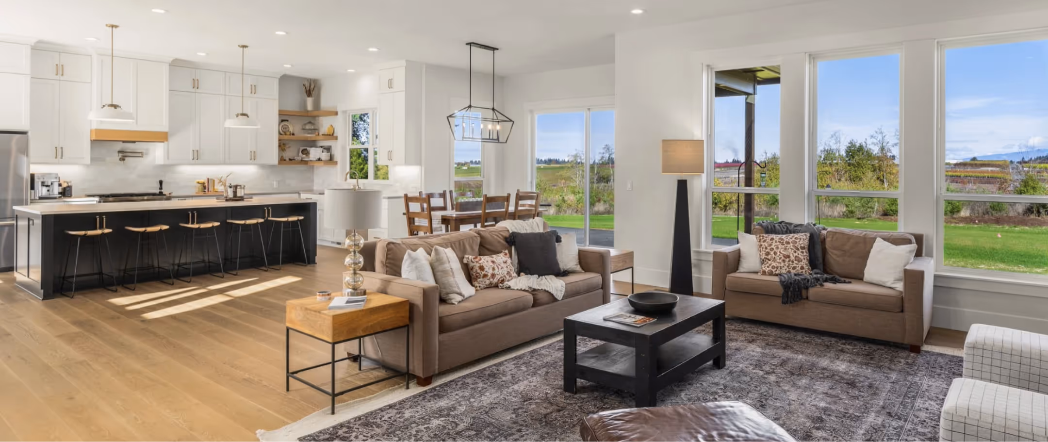 Spacious open-plan living area with modern kitchen, brown sofas, black coffee table, and large windows showing a green outdoor view.