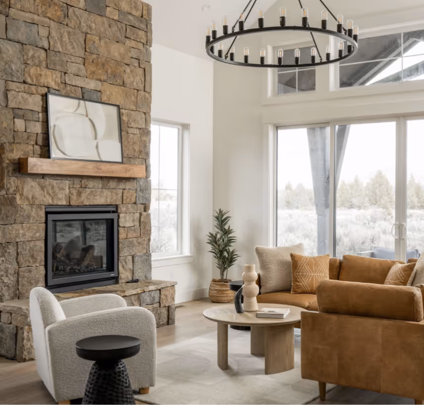 Modern living room with stone fireplace, white armchair, round wooden coffee table, tan sofa, and large windows with outdoor view.