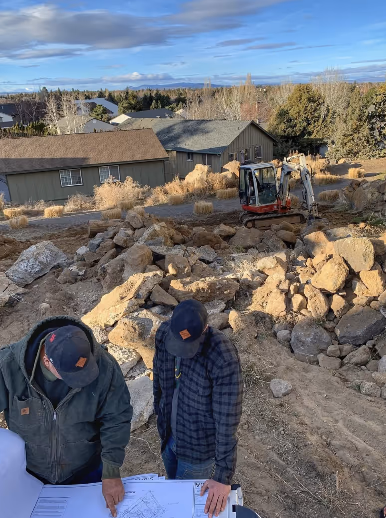 Two men in work jackets and caps reviewing construction blueprints outdoors at a rocky residential site with houses and trees in the background.