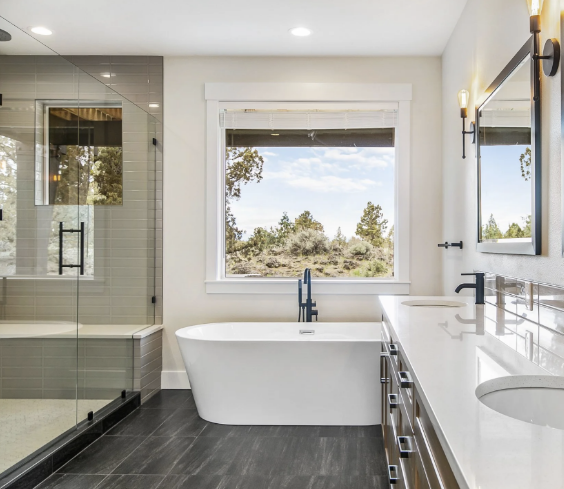 Modern bathroom with freestanding white bathtub under a large window, a glass-enclosed shower, and a double sink vanity with wall-mounted lights.