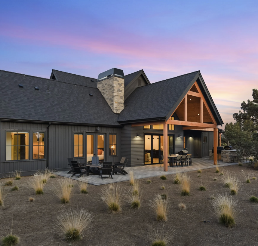 Modern dark gray house with a large patio, wooden beams, outdoor seating area, and evening sky with pink and purple hues.