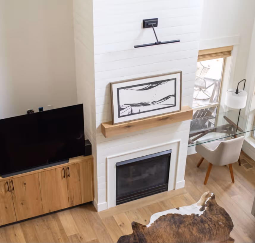 Modern living room with a TV on a wooden cabinet, a white brick fireplace with artwork on the mantel, a glass desk with a white chair, and large windows.