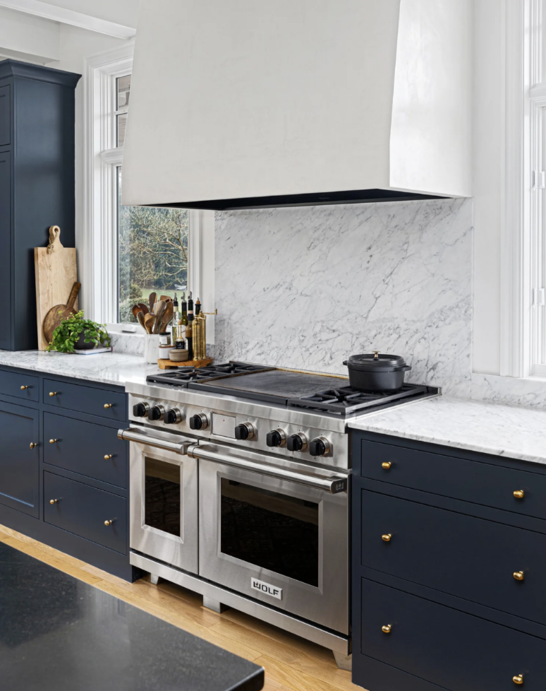Modern kitchen with stainless steel Wolf double oven and stove, navy blue cabinets with gold knobs, white marble countertops and backsplash, and kitchen utensils near the window.