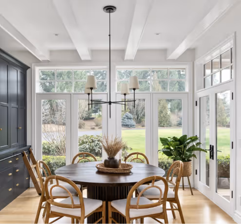 Bright dining room with large windows and glass doors overlooking a garden, featuring a round wooden table surrounded by six cushioned wooden chairs, a modern chandelier, and a potted plant near the door.