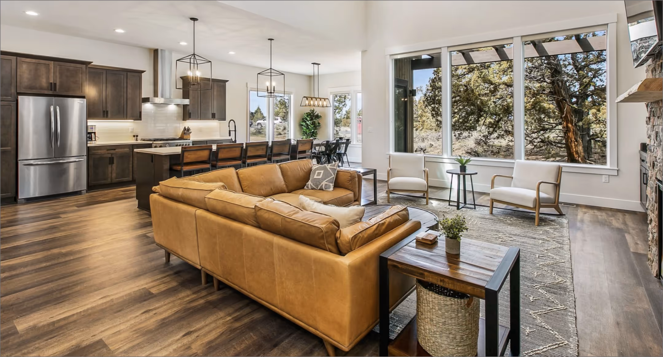 Open-concept living room and kitchen with tan leather sectional sofa, wooden floors, large windows, and modern dark wood cabinetry.
