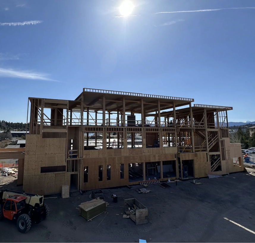 Wooden framing structure of a multi-story building under construction with clear blue sky and sun overhead.