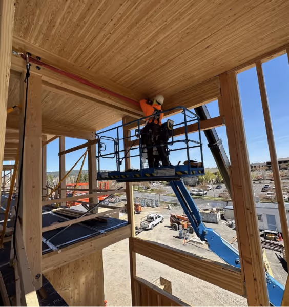 Construction worker in a safety harness on a blue aerial lift working on wooden ceiling beams inside a building frame.