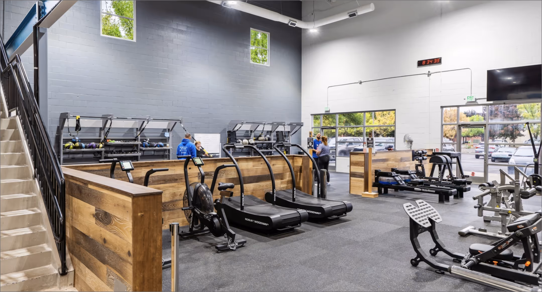 Modern gym interior with cardio and strength equipment, wooden partitions, and people conversing near the entrance.