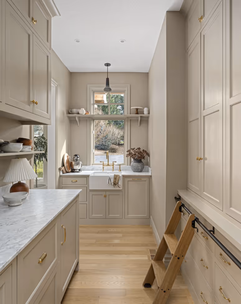 Narrow beige kitchen with marble countertops, gold hardware, wooden floor, farmhouse sink, window, and a wooden step ladder.