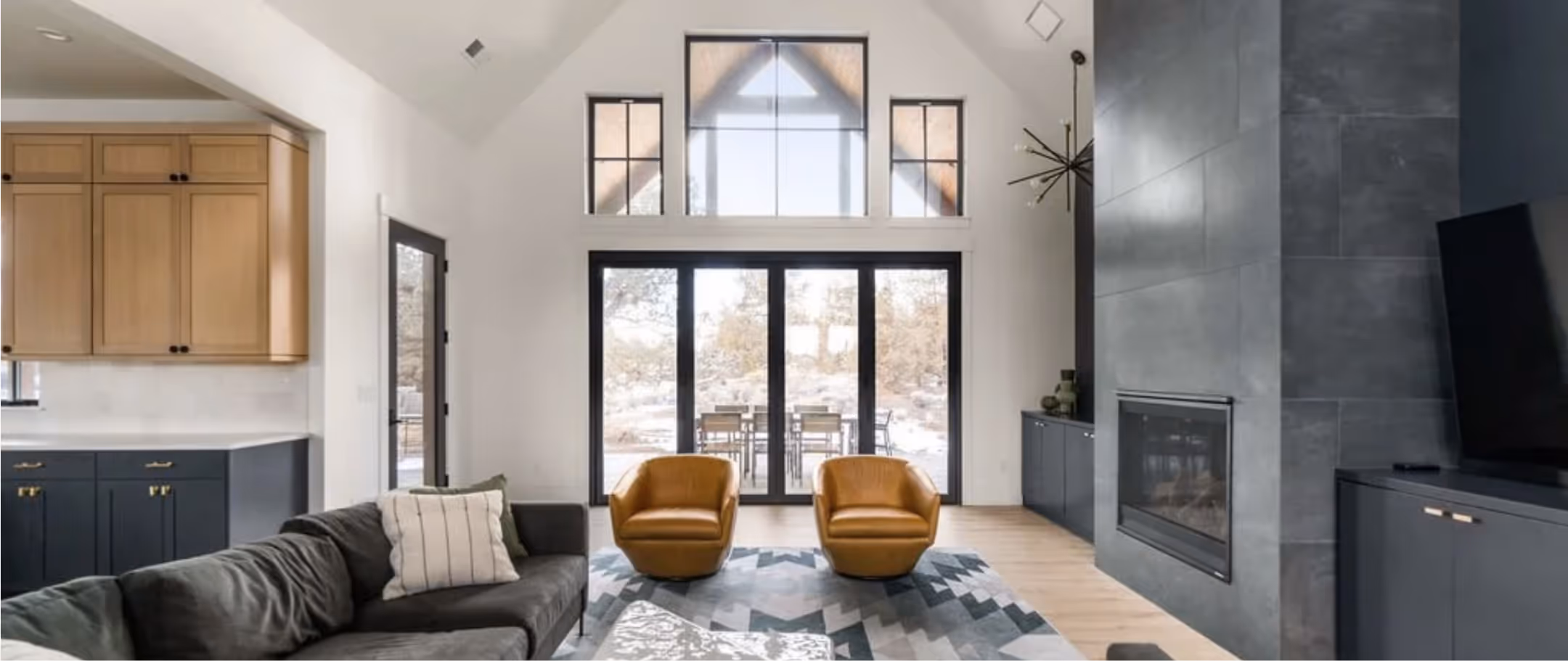 Modern living room with vaulted ceiling, black-framed large windows, two mustard leather chairs, dark gray sofa, geometric rug, and gray tiled fireplace.